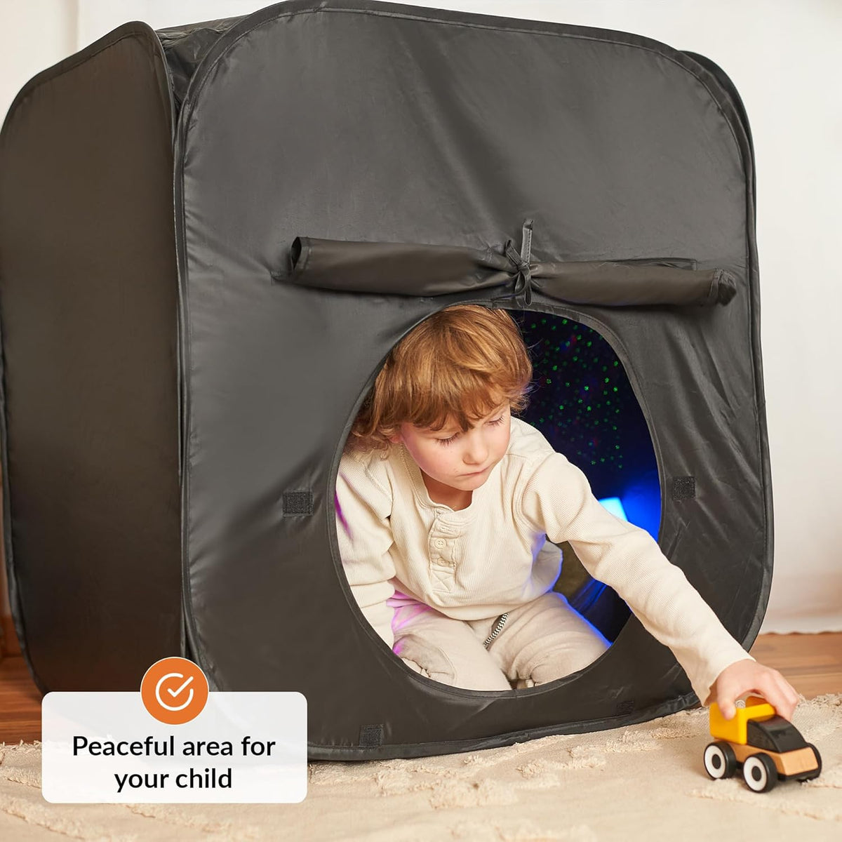 Child playing with a toy car inside a dark play tent with a starry light on a light wooden floor.