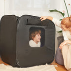 Black play tent with a child's face peeking out, placed on a wooden floor with a mom sitting nearby.