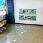 Children's classroom with a sequins board displaying 'MOM ❤️ HOME' and educational materials on shelves.