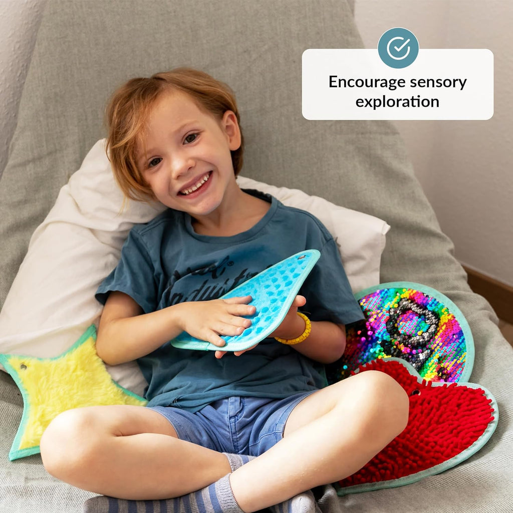 Child holding a blue textured toy with colorful toys around, sitting on a cushion.
