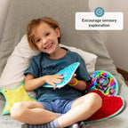 Child holding a blue textured toy with colorful toys around, sitting on a cushion.