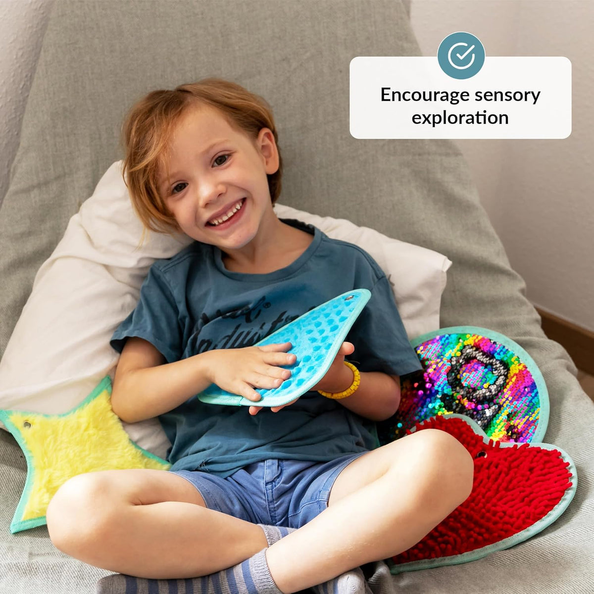 Child holding a blue textured toy with colorful toys around, sitting on a cushion.