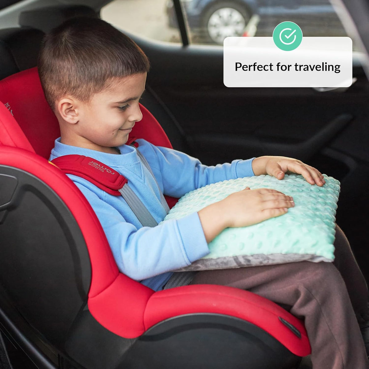 Child sitting in a red car seat holding a green pillow with text 'Perfect for traveling'.