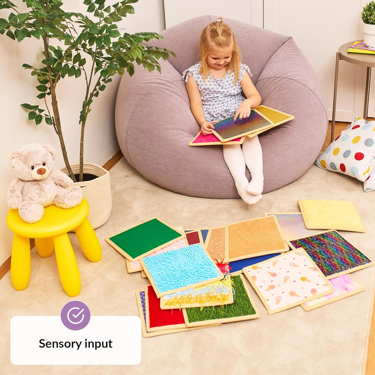 Child sitting on a bean bag touching sensory sheets with educational toys around